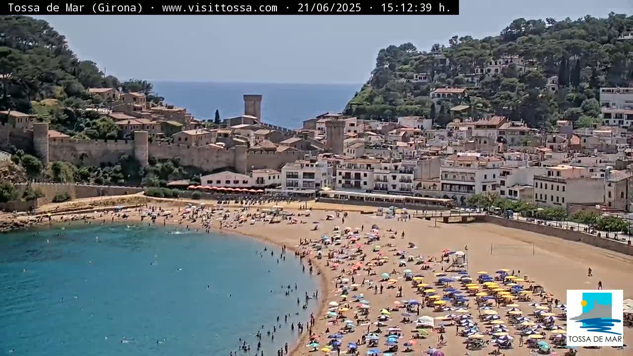 A crowded beach curves around a calm bay, in front of a town nestled against a hillside on a sunny day.