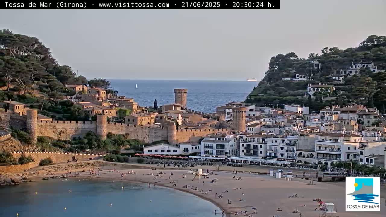 A coastal town with a medieval walled section, a sandy beach with people sunbathing, and calm sea under a clear, light-grey sky.