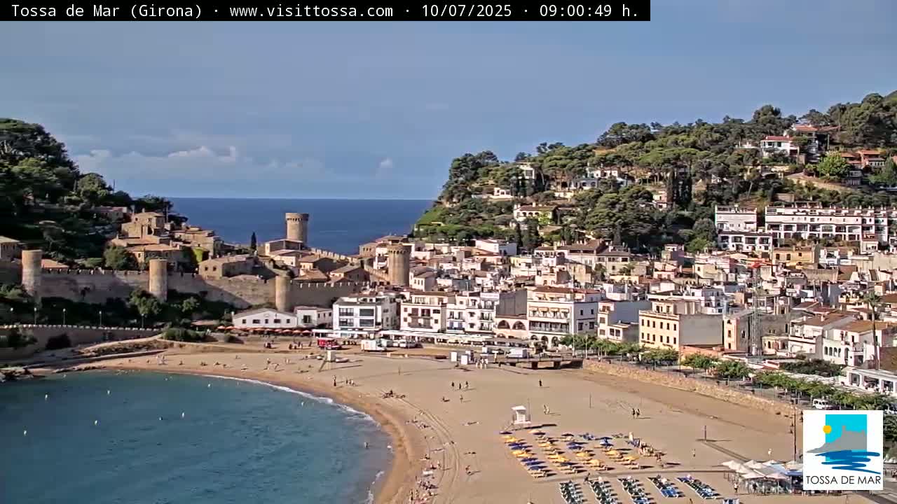 A coastal town with a medieval-style castle sits on a hill overlooking a sandy beach and calm blue water under a mostly clear sky.