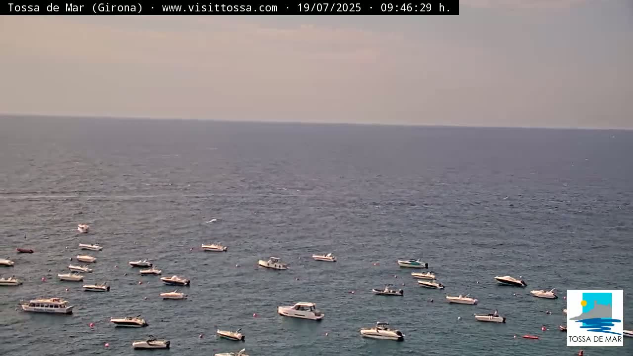 Numerous small boats are anchored in a calm, light-blue sea under a hazy sky.
