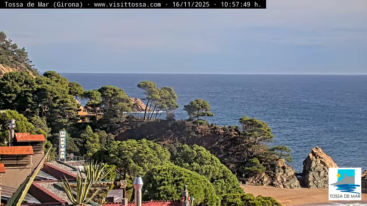 A sunny day reveals a clear blue sea bordered by lush green, pine-covered hills descending to rocky shores and a small sandy beach, with rooftops visible in the foreground.