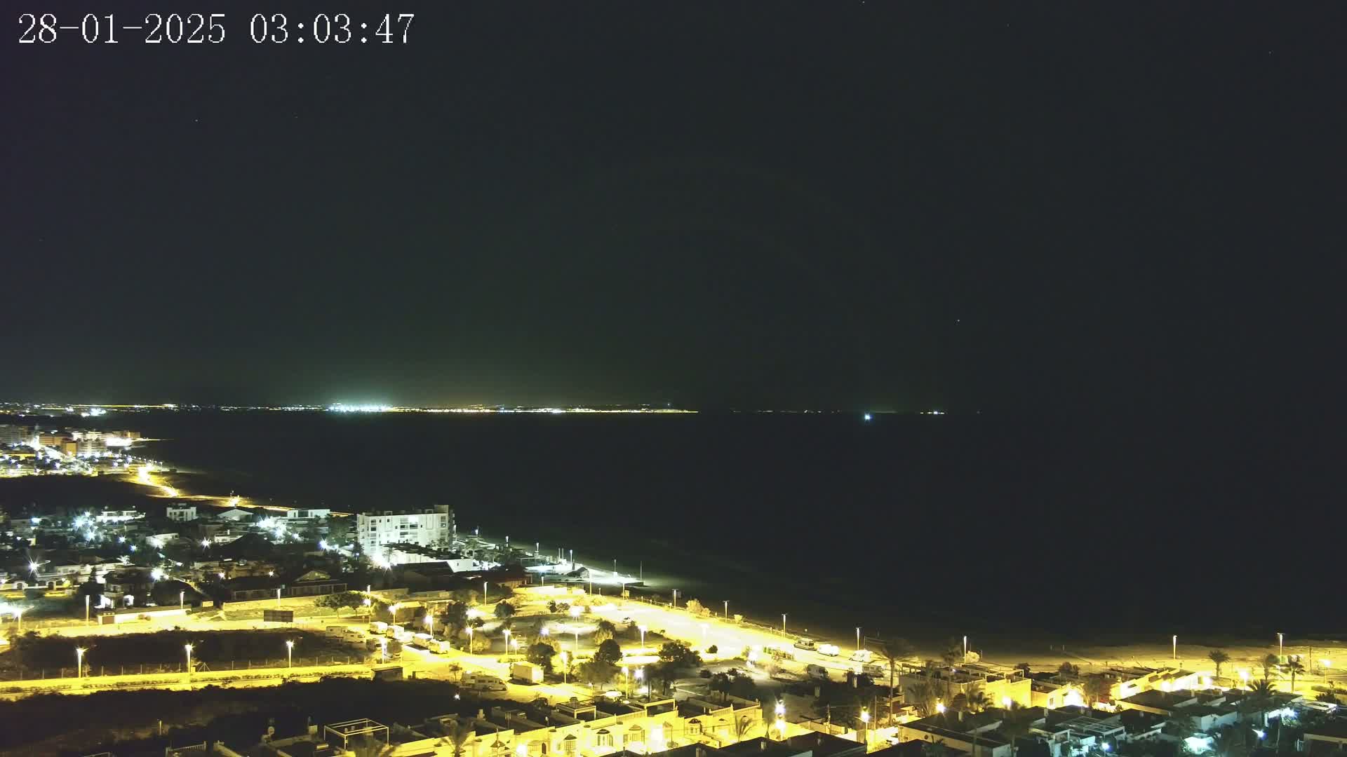 A nighttime coastal scene shows a dark ocean, a dimly lit beach, and a residential area illuminated by streetlights under a clear, starry sky.