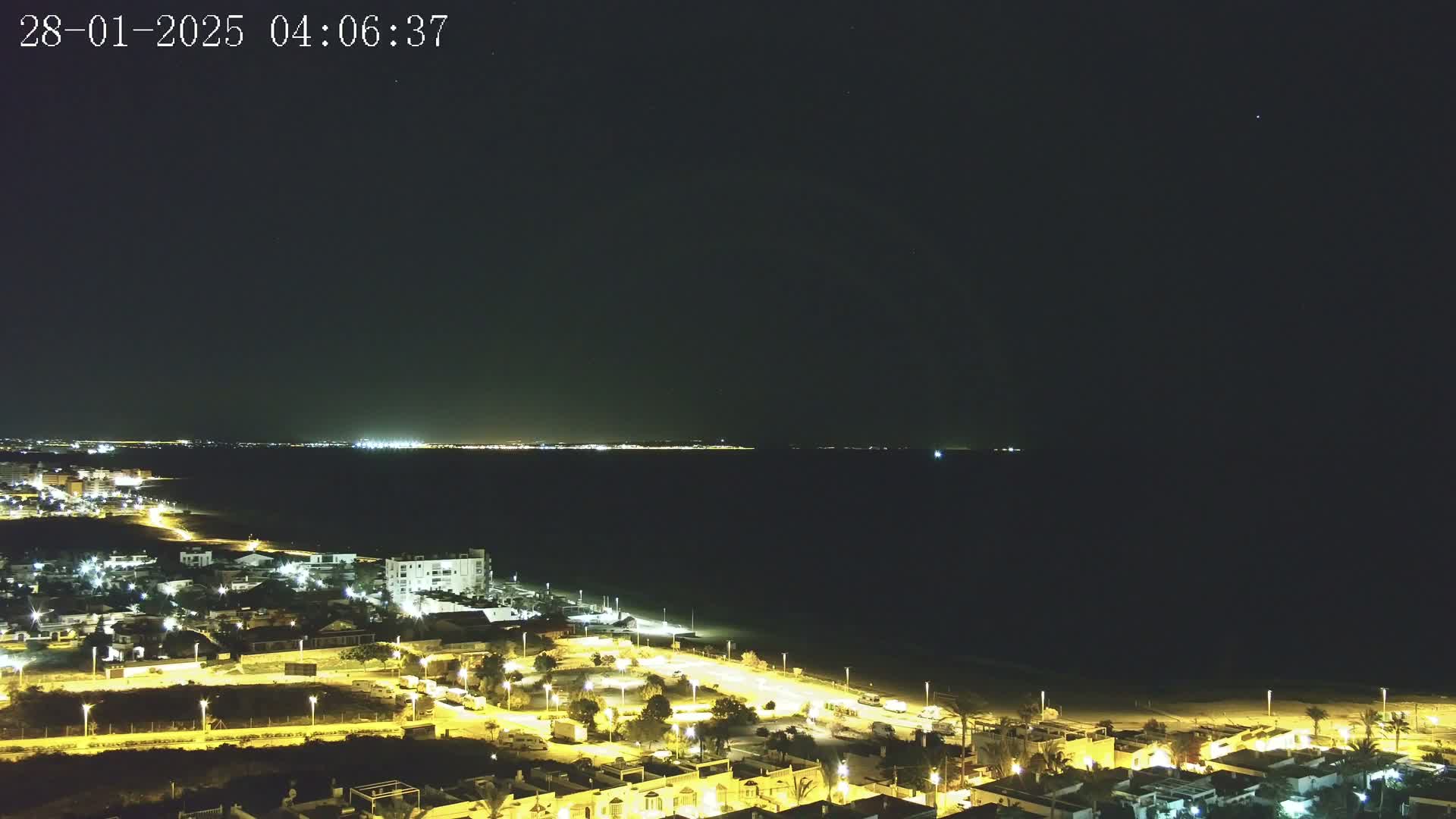 A nighttime coastal scene shows a dark ocean with a faintly lit shoreline and a town illuminated by streetlights under a clear, dark sky.