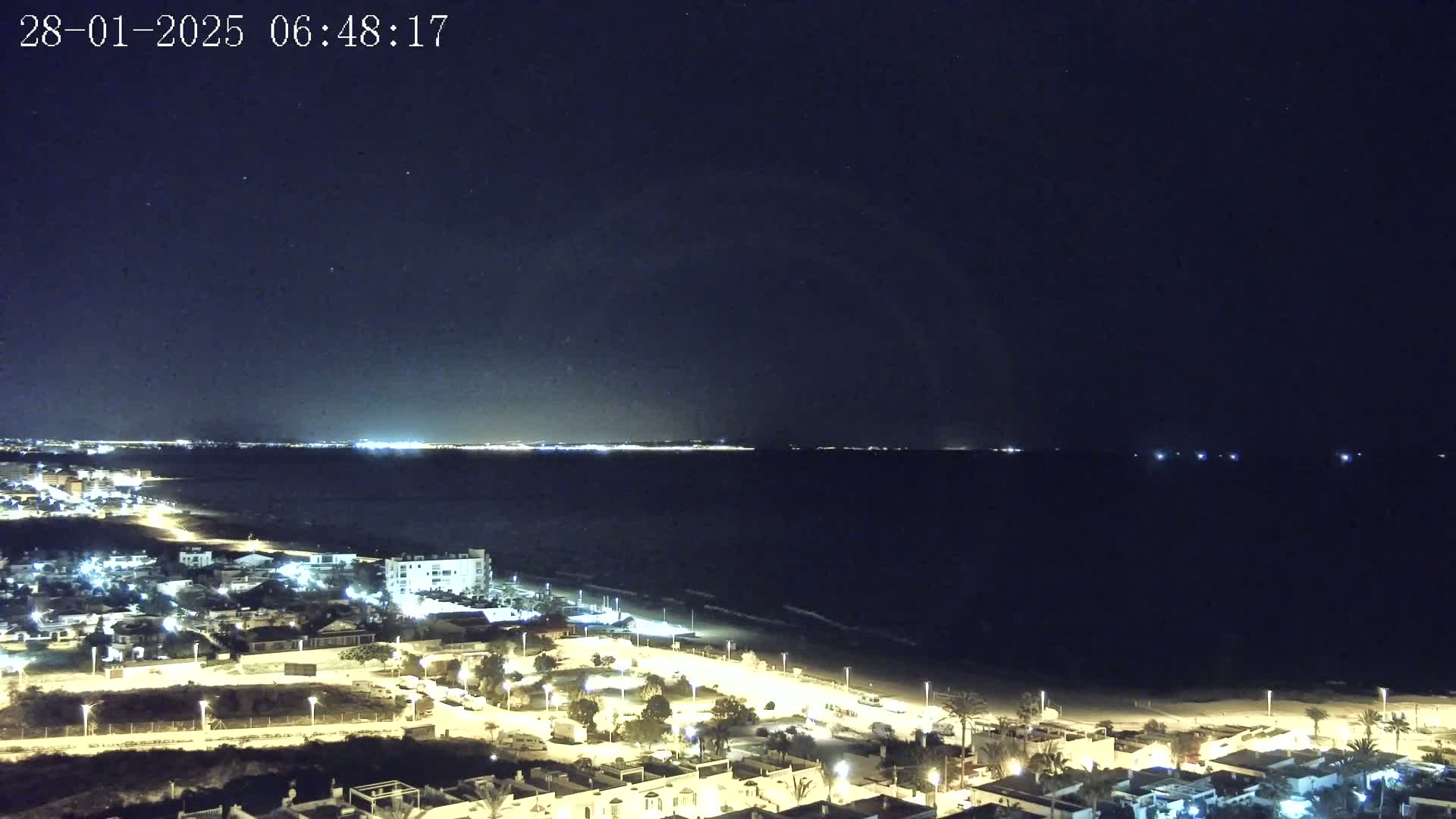 A nighttime coastal scene shows a dark ocean under a clear, starry sky, with a brightly lit town and beach visible in the foreground.