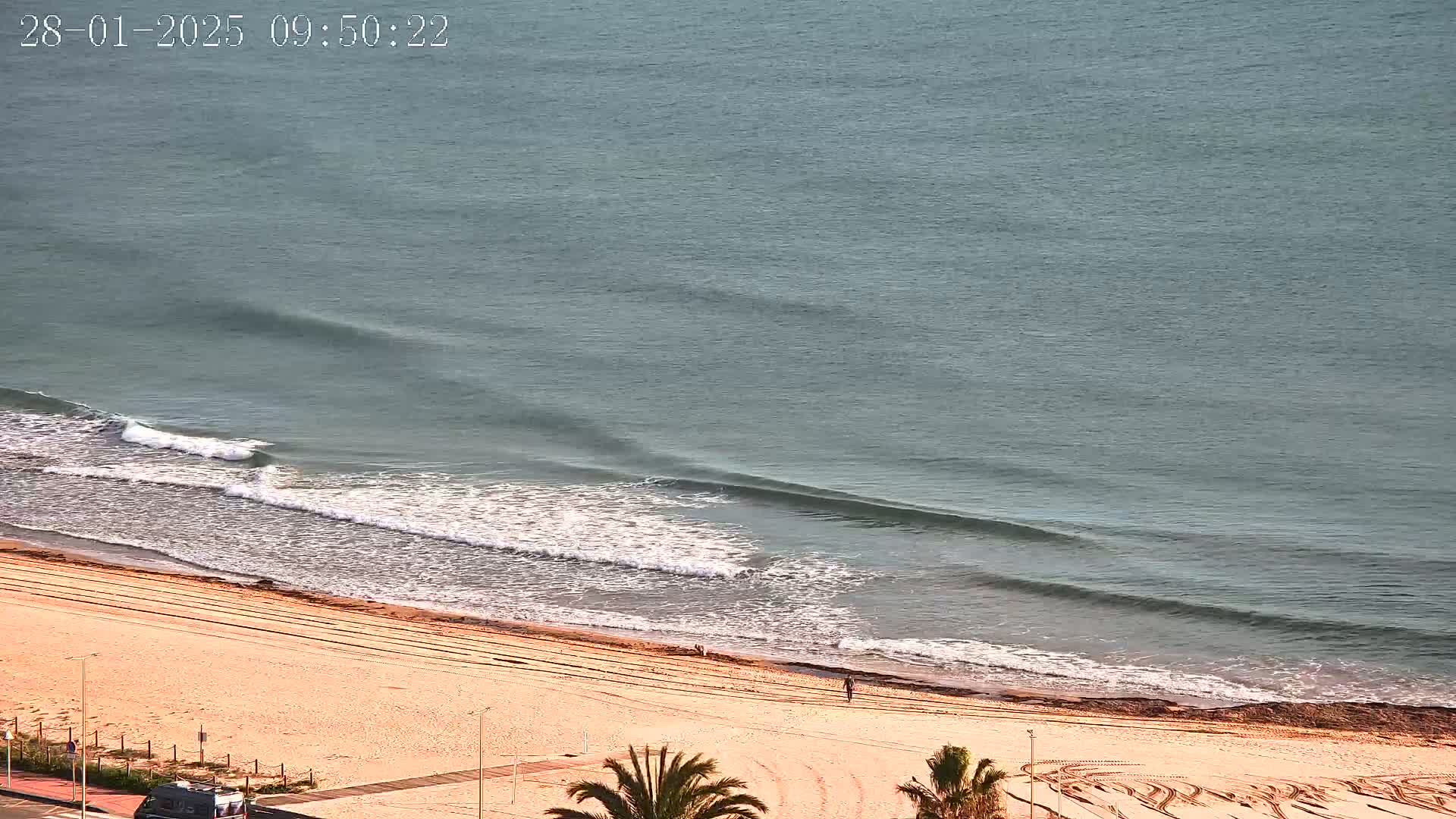 A sandy beach with tire tracks and a lone person walking on it is met by gently rolling waves on a calm sea.