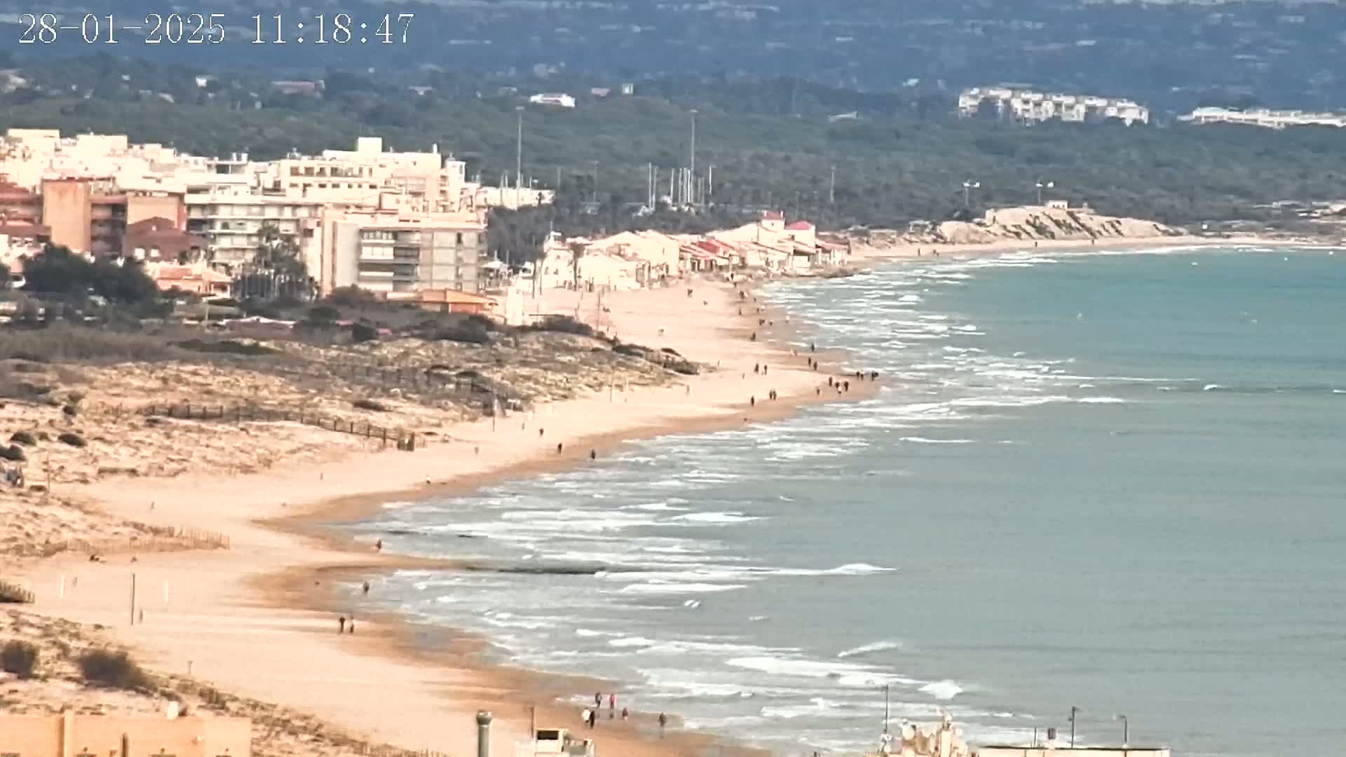 A long sandy beach with scattered people stretches along a calm, light-blue sea under a mostly cloudy sky, backed by buildings and low hills.
