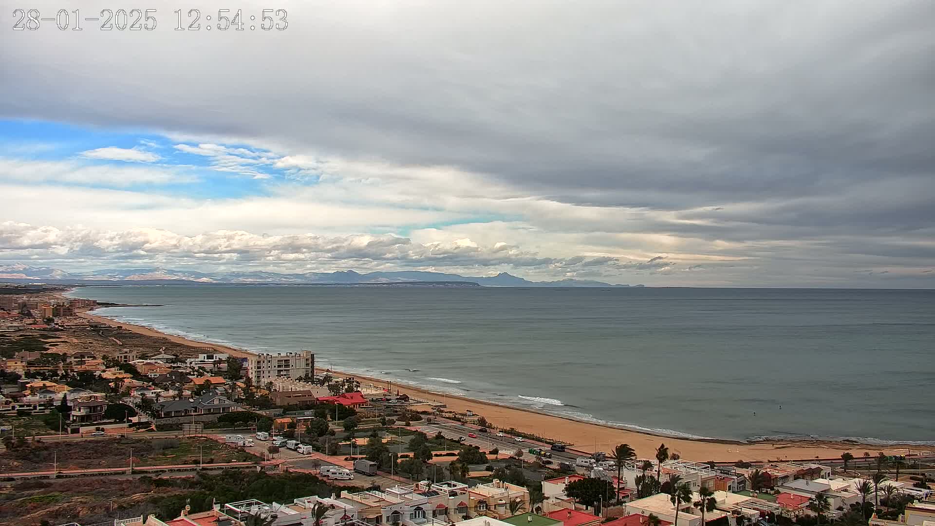 A coastal town overlooks a wide sandy beach and calm ocean under a partly cloudy sky with dark clouds dominating the horizon and distant mountains visible in the background.