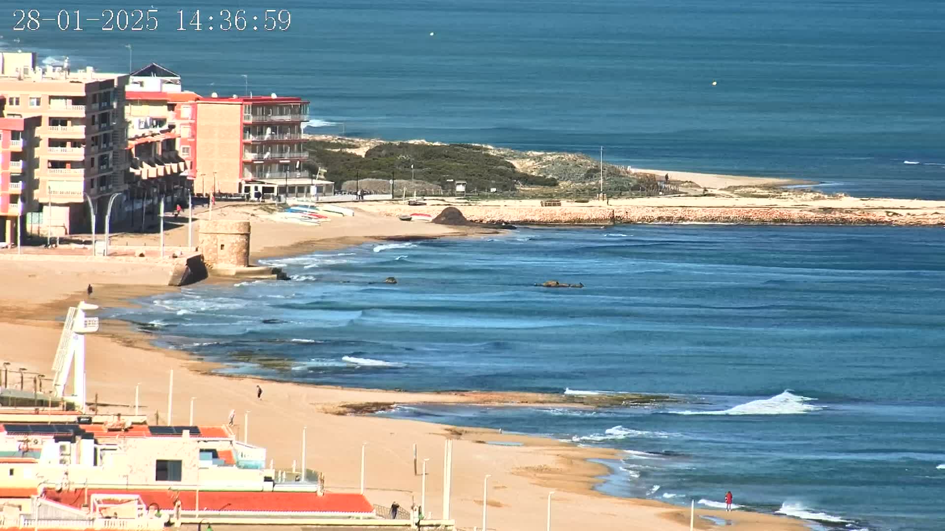 A sandy beach with a few people on it, buildings, and a small stone tower next to calm, blue ocean waves under a sunny sky.