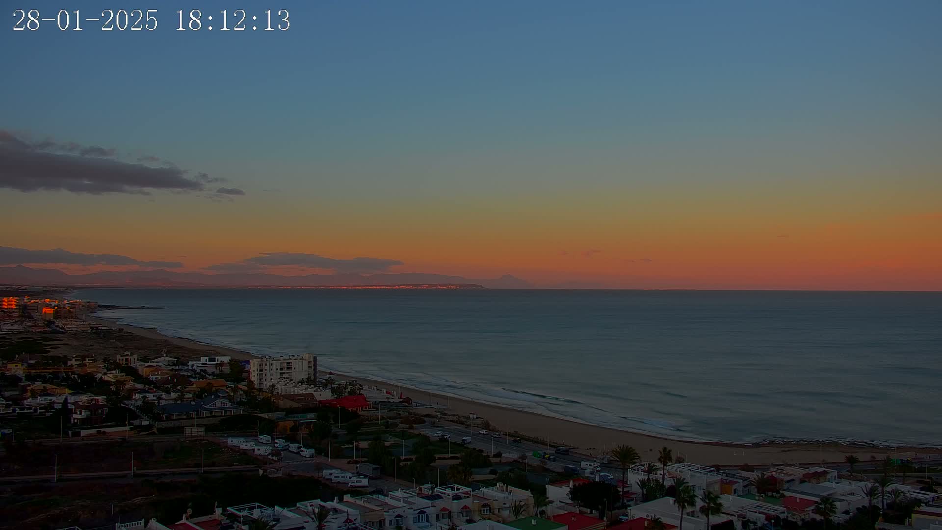 A coastal town at sunset overlooks a calm ocean with a distant mountain range under a mostly clear, orange and pink sky.