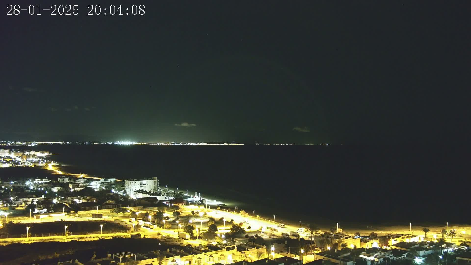 A nighttime coastal scene shows a dark body of water with faintly lit far shores, and a nearby town illuminated by streetlights.