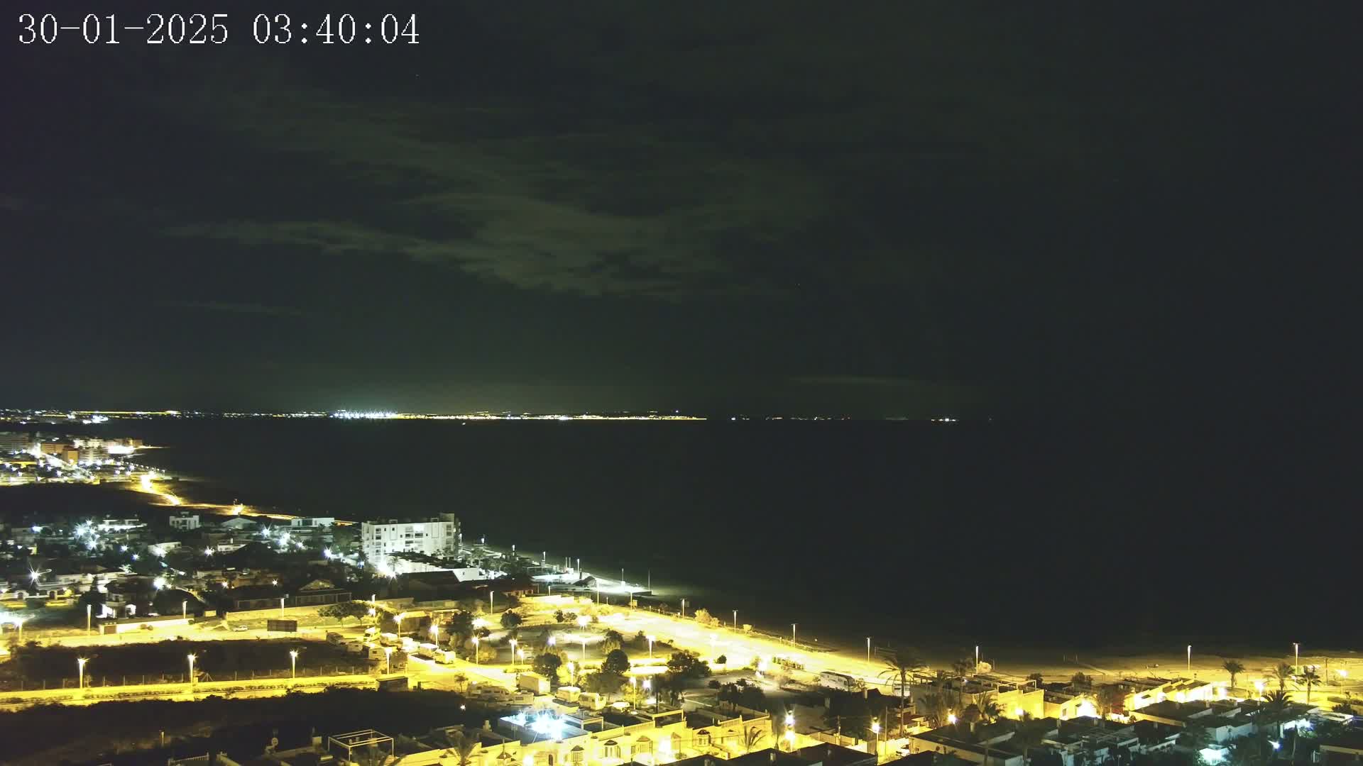 A nighttime coastal town illuminated by streetlights overlooks a dark, calm sea under a mostly cloudy sky.