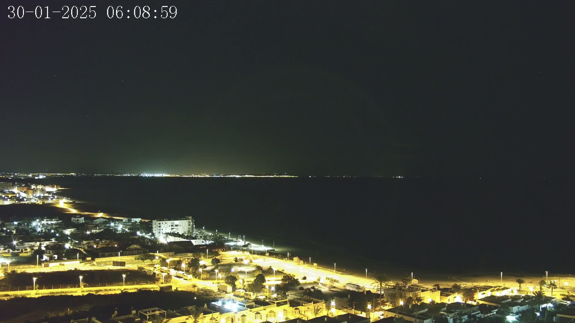 A nighttime, aerial view of a coastal town with its lights illuminating the streets and buildings, a dark beach, and a calm, dark sea extending to a distant, faintly lit coastline under a clear night sky.