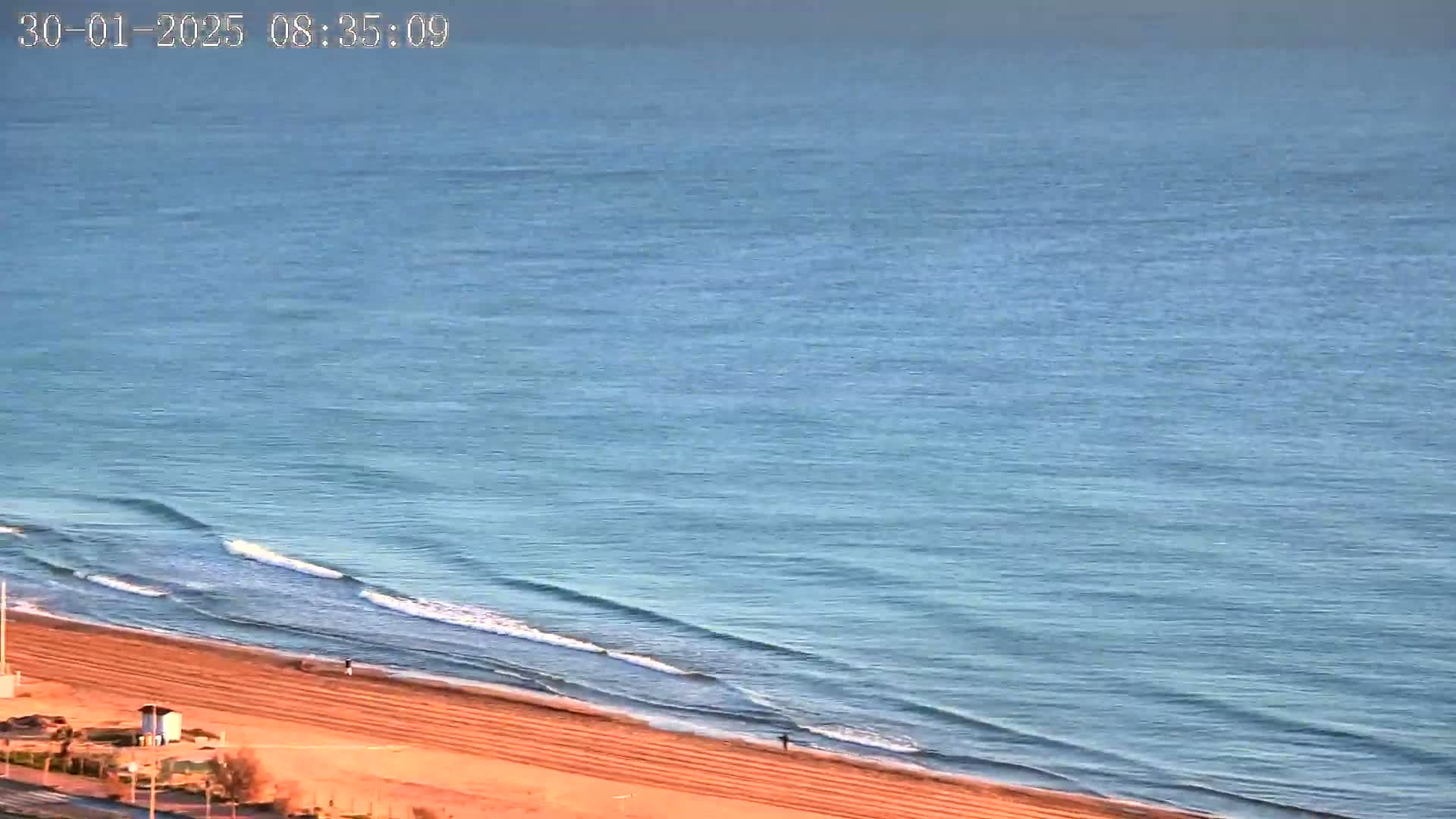 A mostly calm ocean with small waves rolls onto a sandy beach under a clear sky.