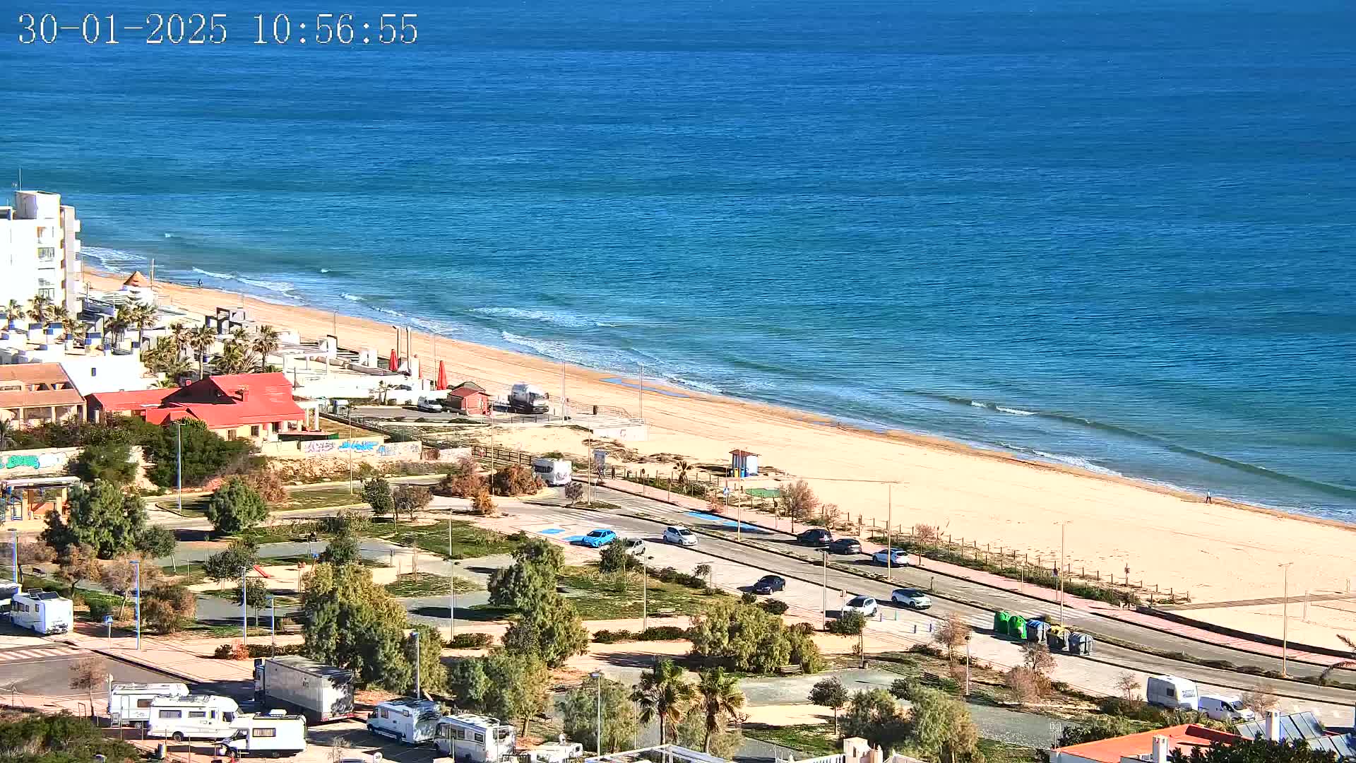 A sandy beach with gentle waves and blue water is adjacent to a coastal road lined with parked cars and recreational vehicles, with buildings and greenery inland on a sunny day.