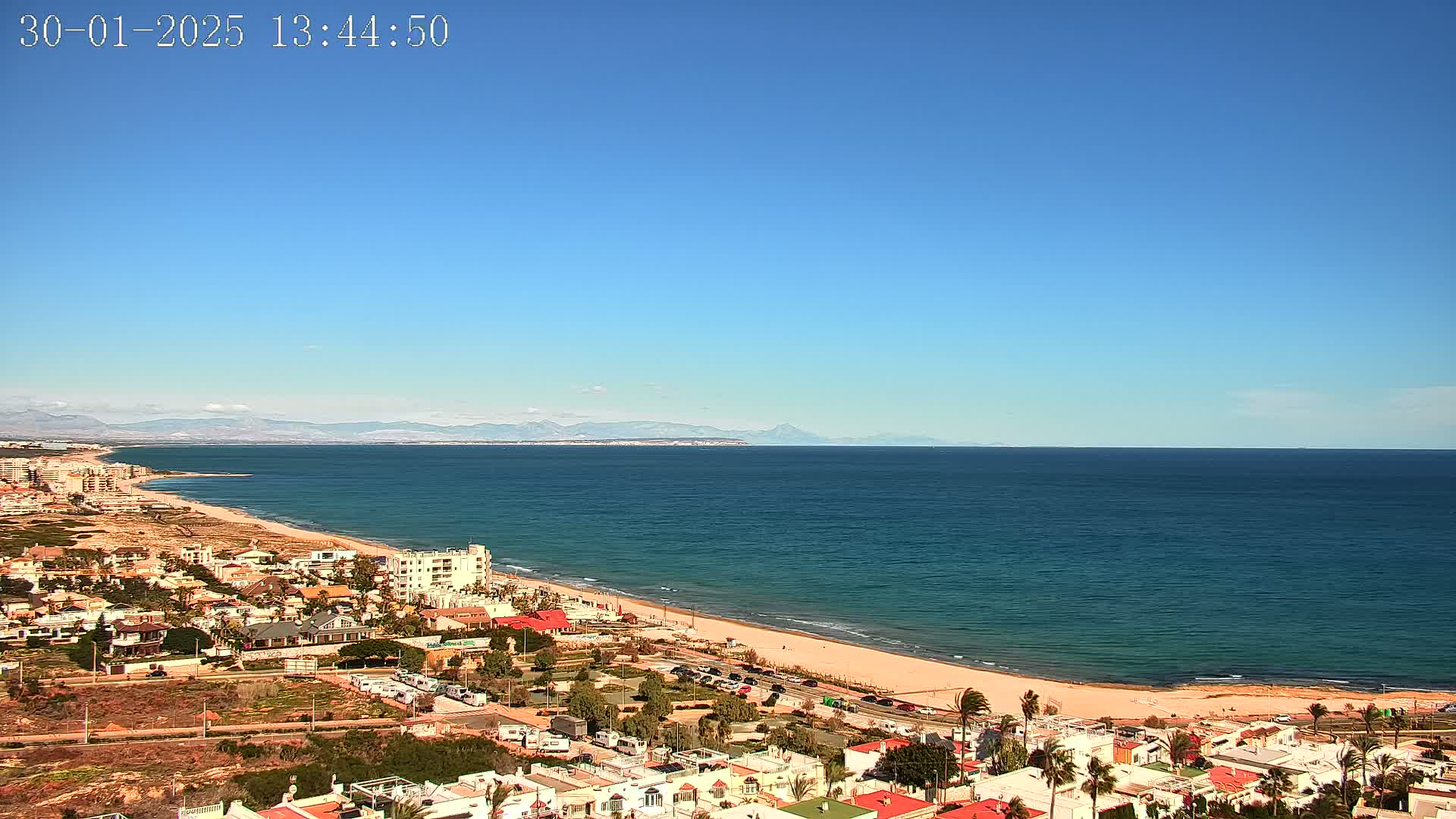 A coastal town overlooks a calm, blue ocean stretching to distant mountains under a clear, sunny sky.