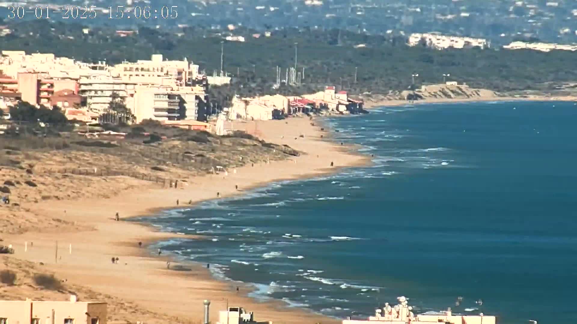 A long sandy beach with a few people walking along it meets a blue ocean with small waves under a sunny sky.