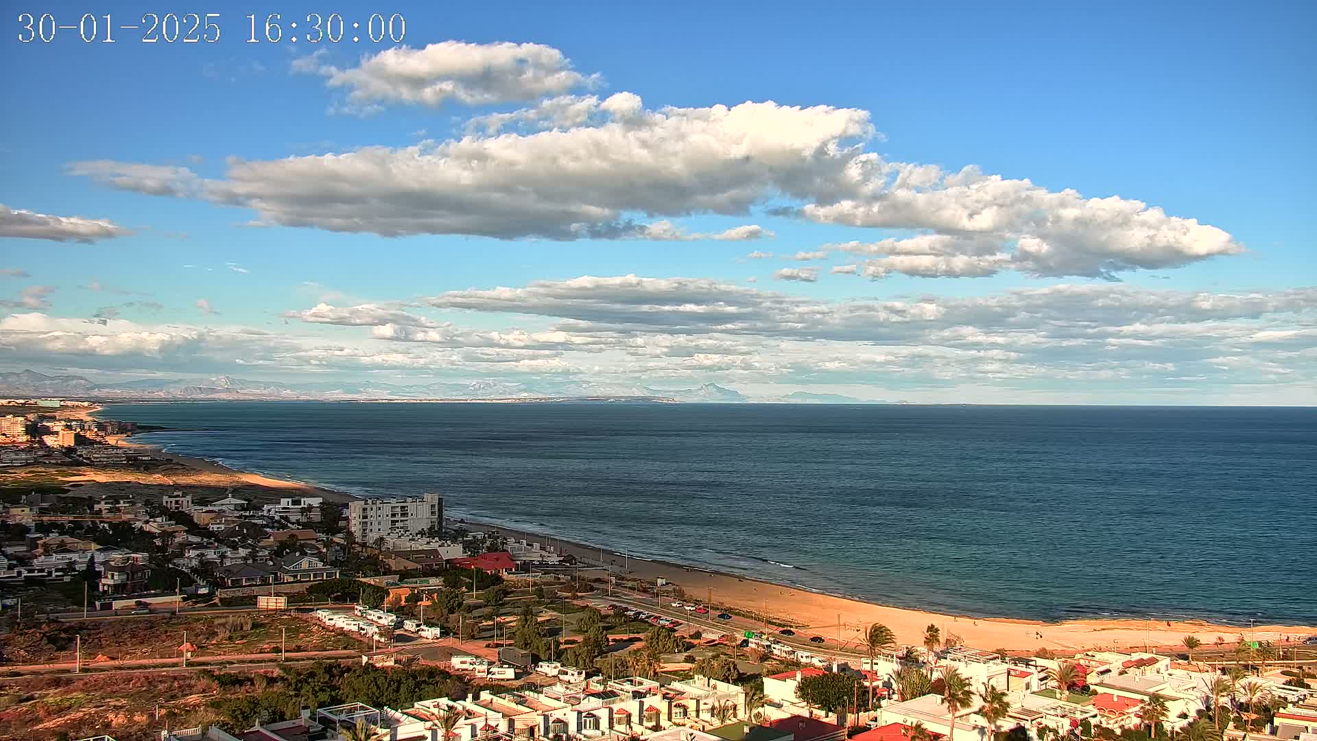 An aerial view of a coastal town on a partly cloudy day shows a sandy beach curving along a dark-blue ocean toward distant mountains.