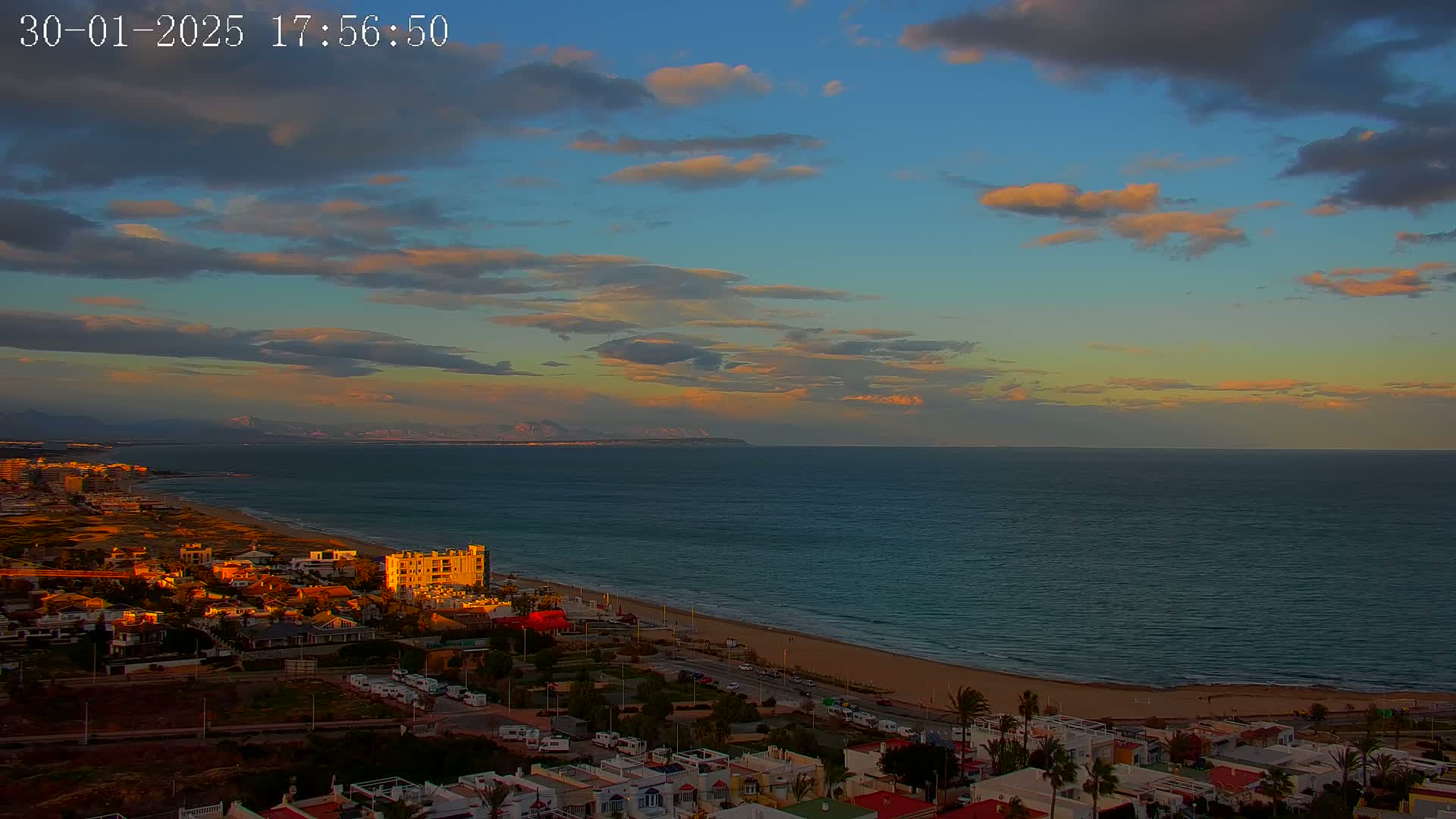 A coastal town at sunset, with a calm sea stretching to distant mountains under a partly cloudy sky.