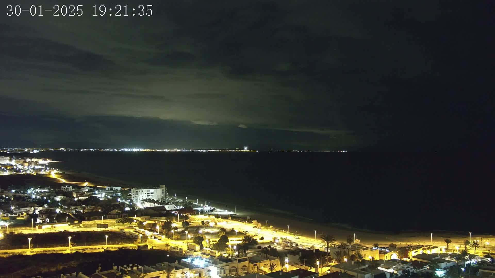 A nighttime coastal town illuminated with streetlights overlooks a dark ocean under a cloudy sky.