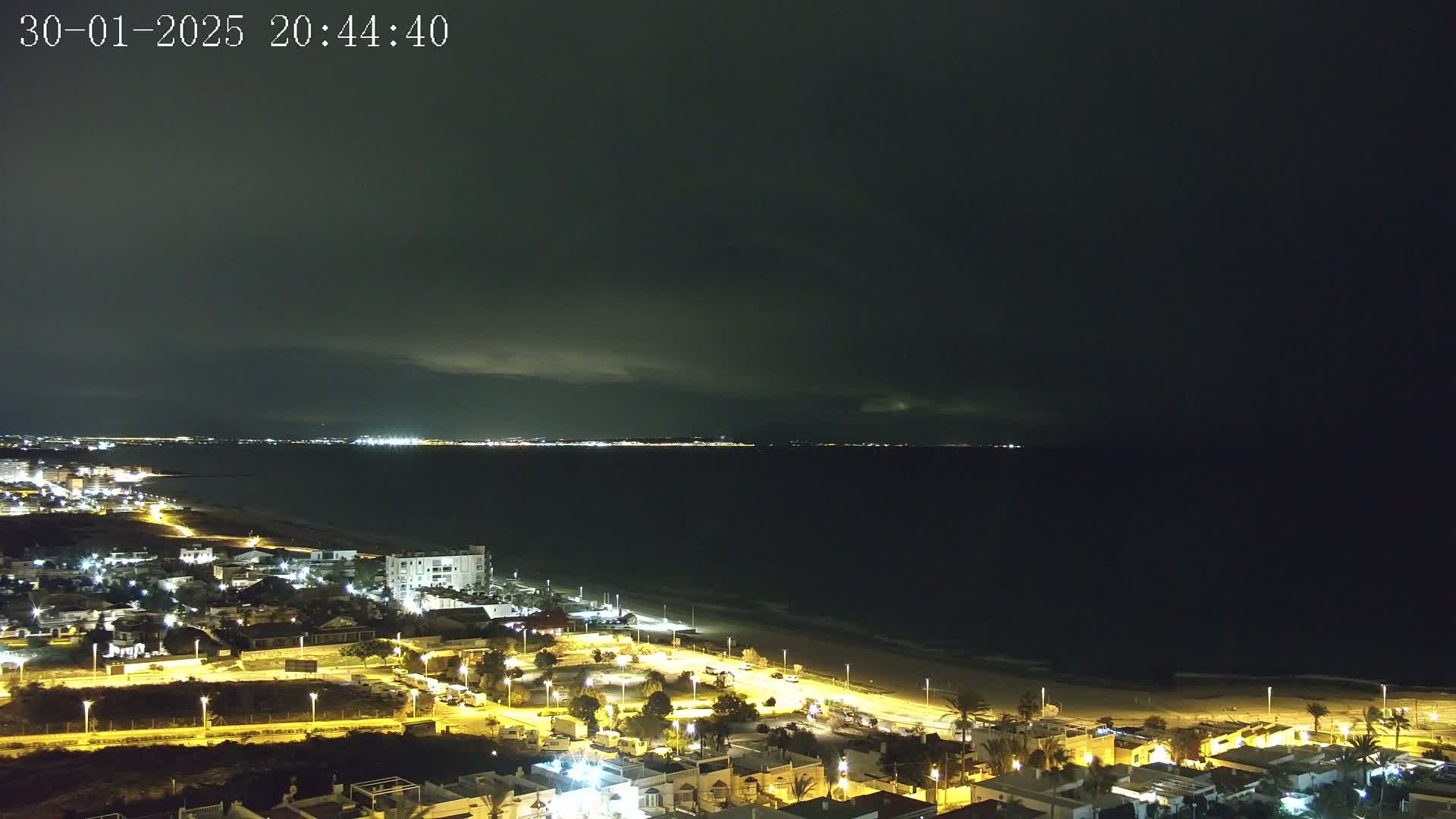 A nighttime coastal scene shows a dark sea under a mostly cloudy sky, with a brightly lit town and beach visible in the foreground.