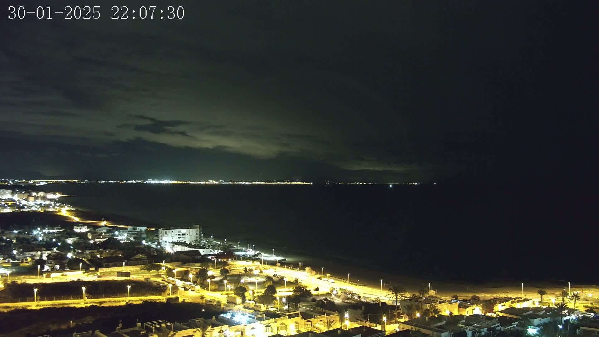 A nighttime coastal town is illuminated, with a dark, cloudy sky over a calm, dark sea.
