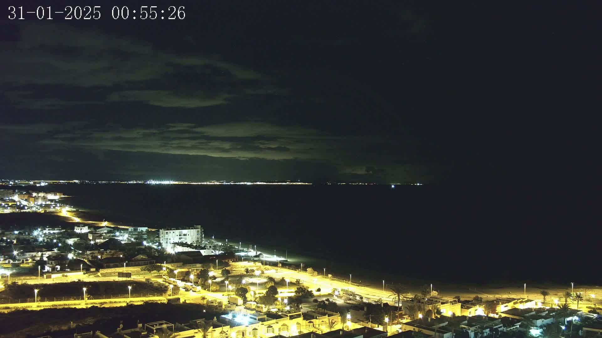 A nighttime coastal town illuminated by streetlights, with a dark sea and a cloudy sky in the background.