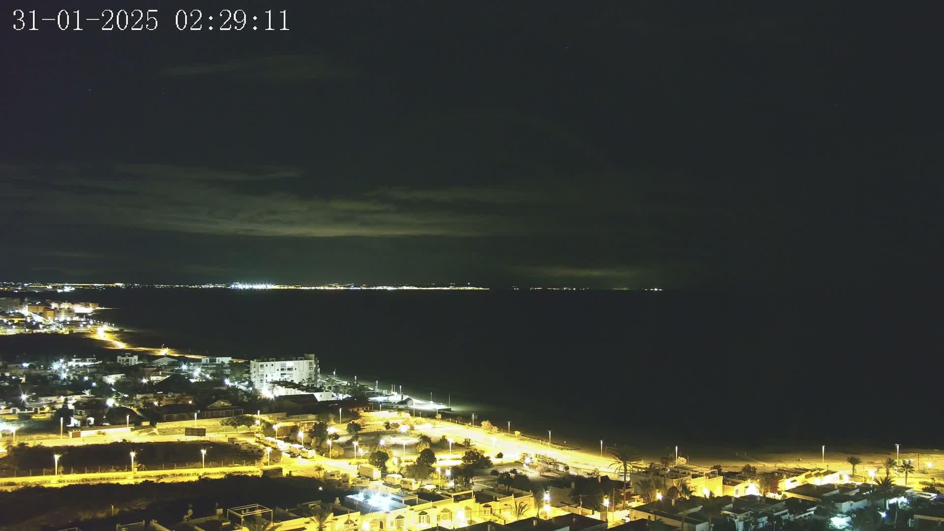 A nighttime coastal scene shows a dark sea under a slightly cloudy sky, with a brightly lit town stretching along the shore.