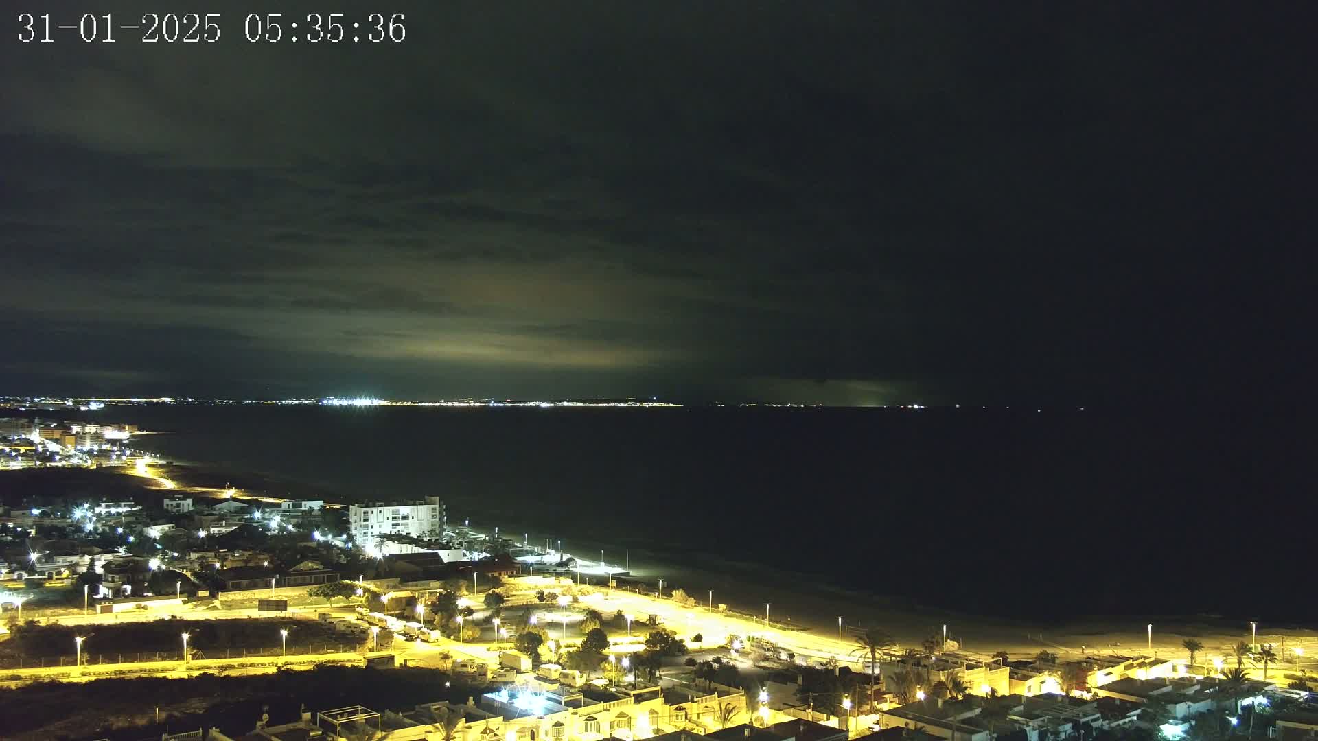 A coastal town at night, illuminated by streetlights, overlooks a dark sea under a mostly cloudy sky.