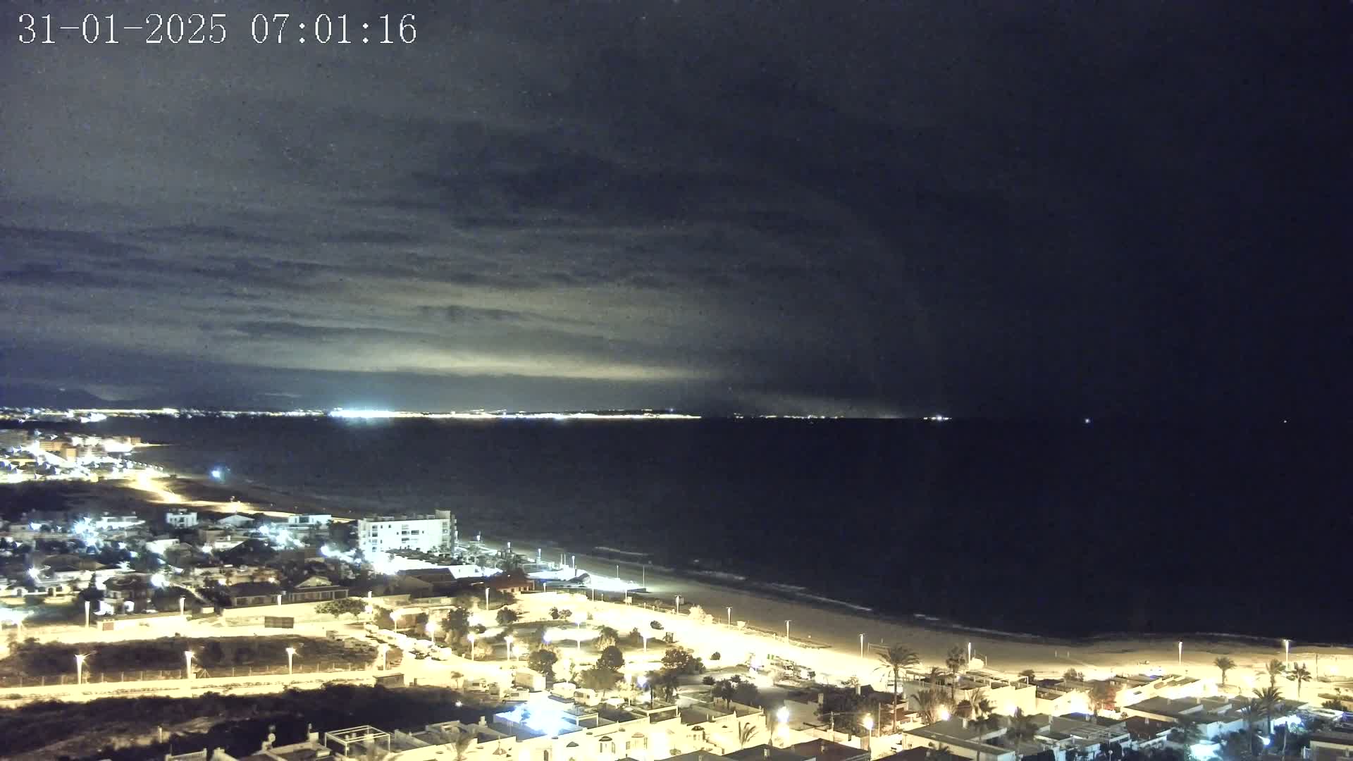 A nighttime coastal scene shows a dark ocean under a cloudy sky, with a brightly lit beachfront town visible in the foreground.