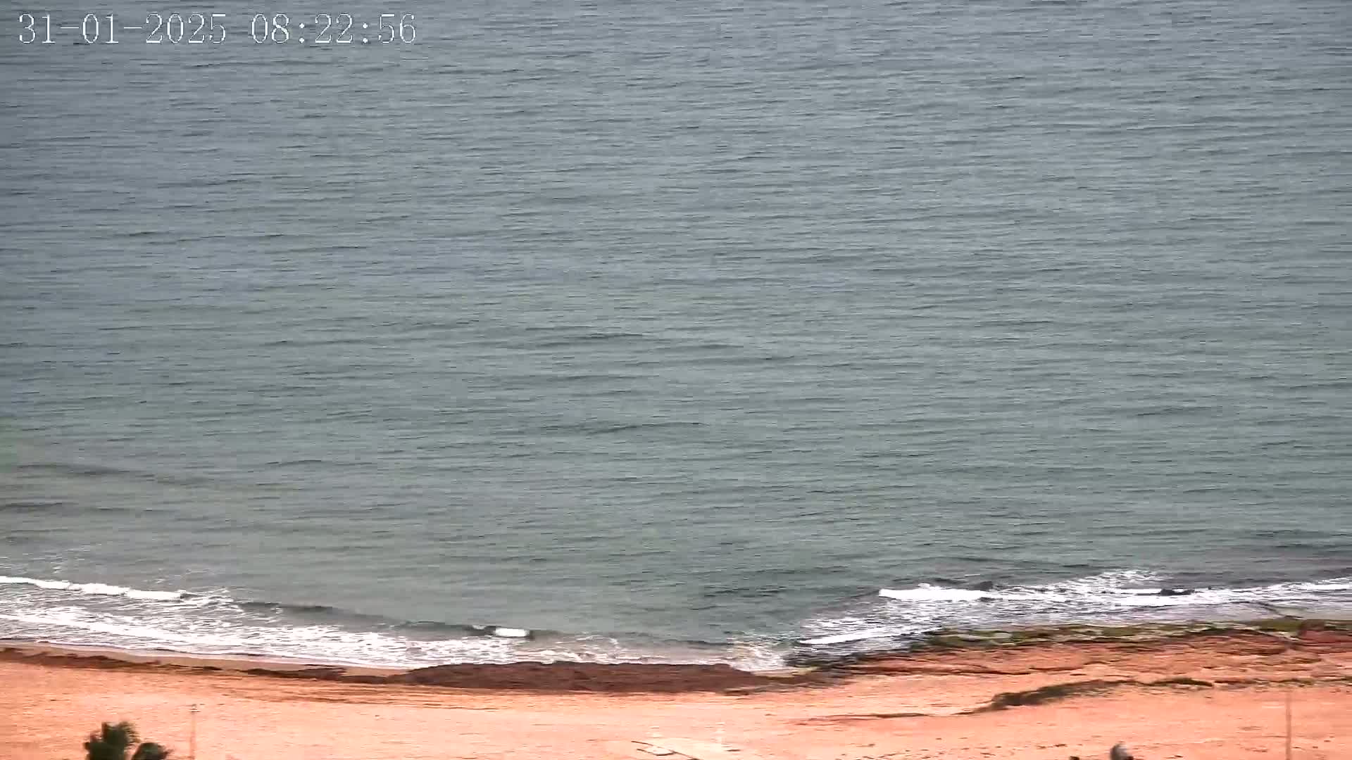 A mostly calm, grey-green sea laps a sandy beach with reddish-brown rocks at the waterline.