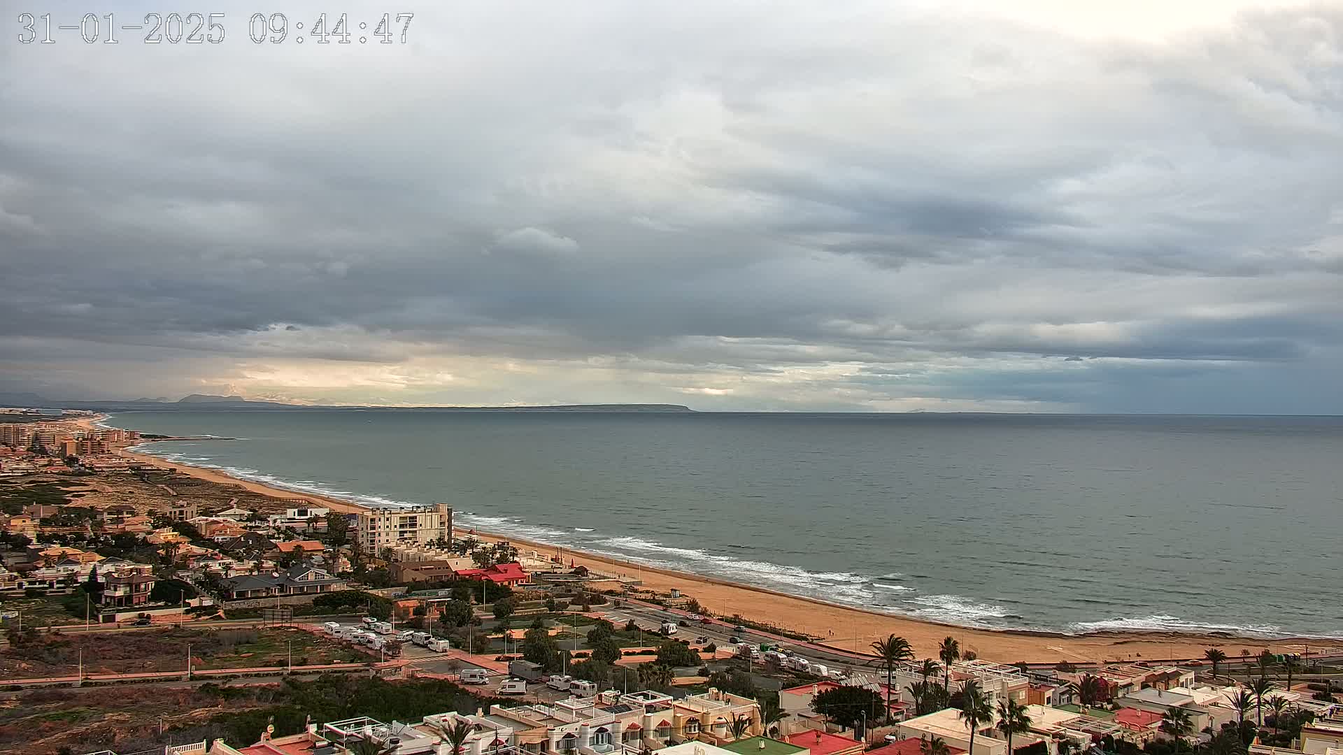 A coastal town with overcast skies overlooks a long sandy beach and calm ocean, with distant mountains barely visible on the horizon.
