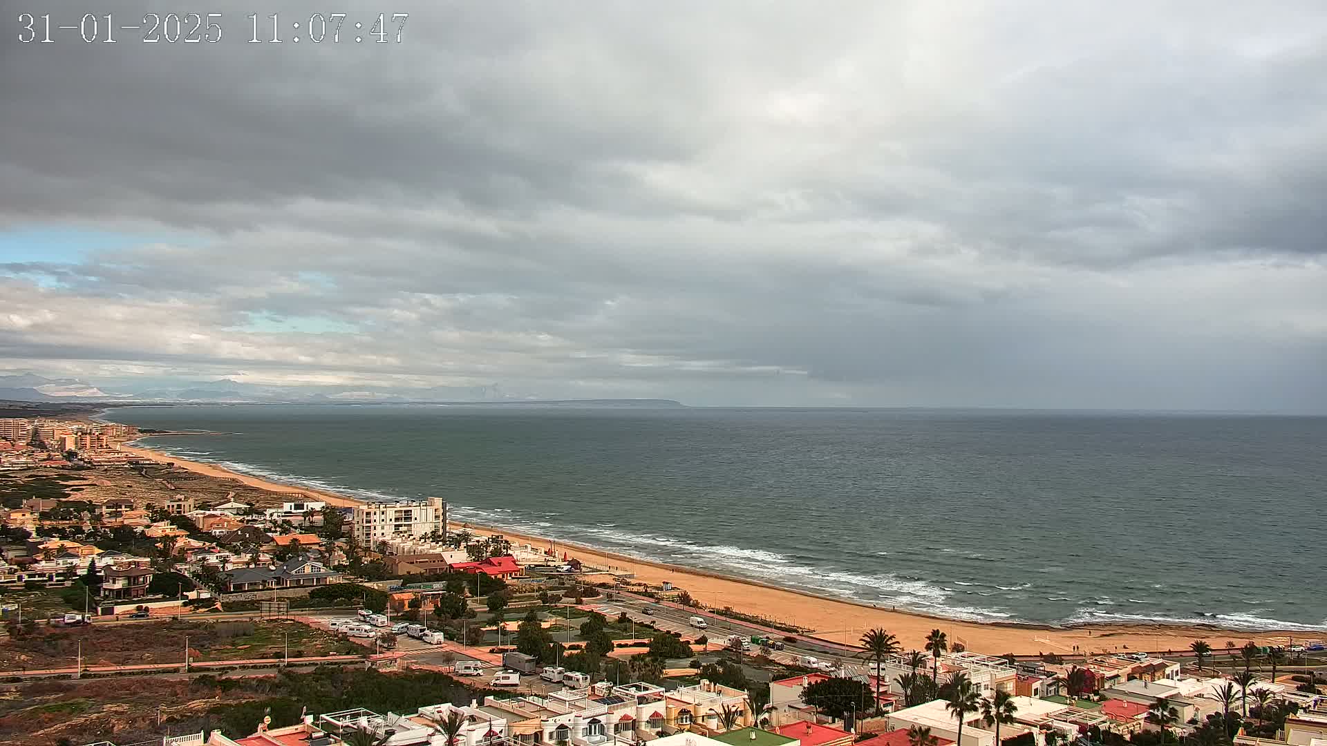 A coastal town with low-rise buildings stretches along a sandy beach and ocean under a mostly cloudy sky.