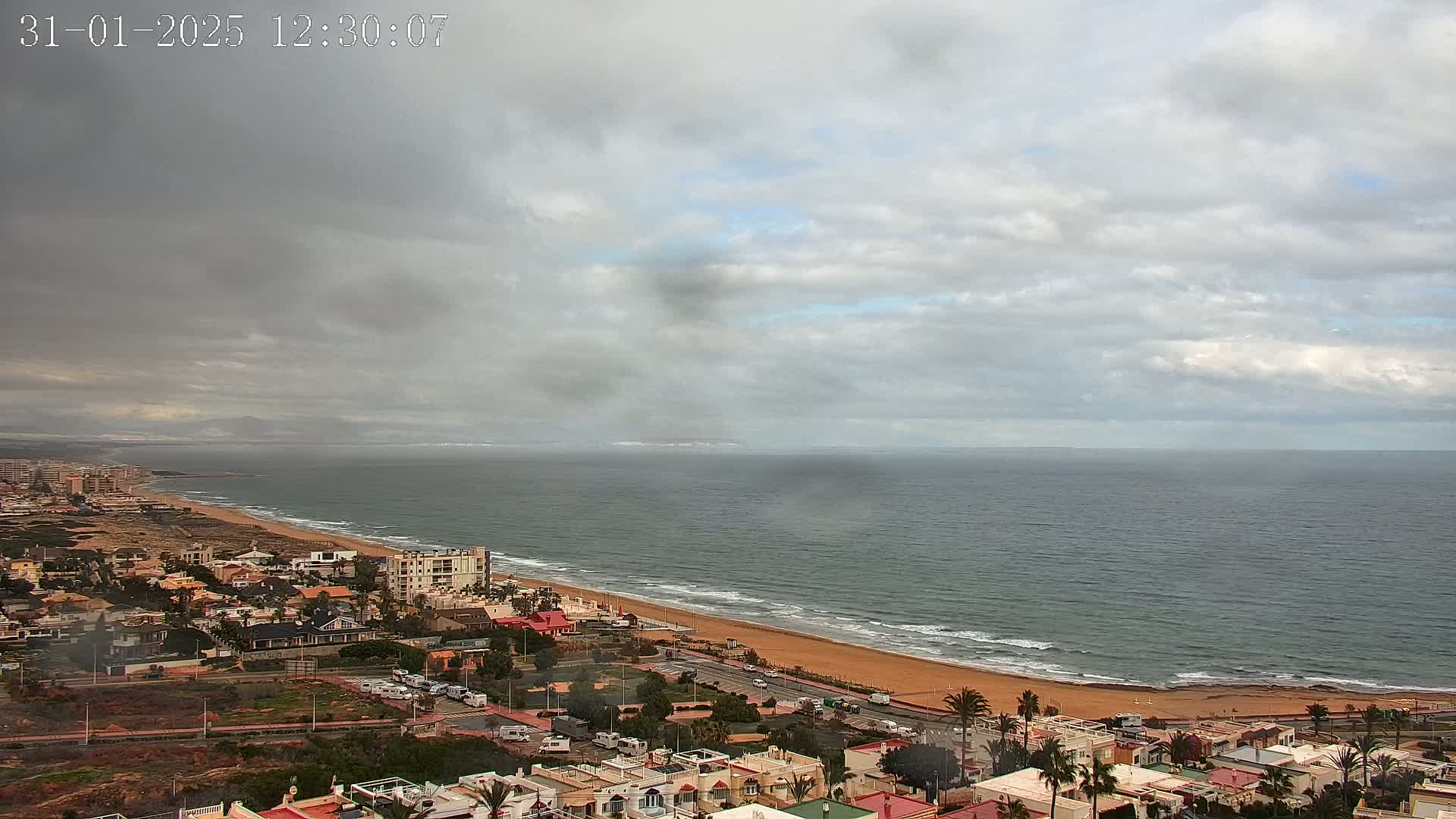 An overcast sky hangs over a coastal town with a long sandy beach and calm ocean waves.