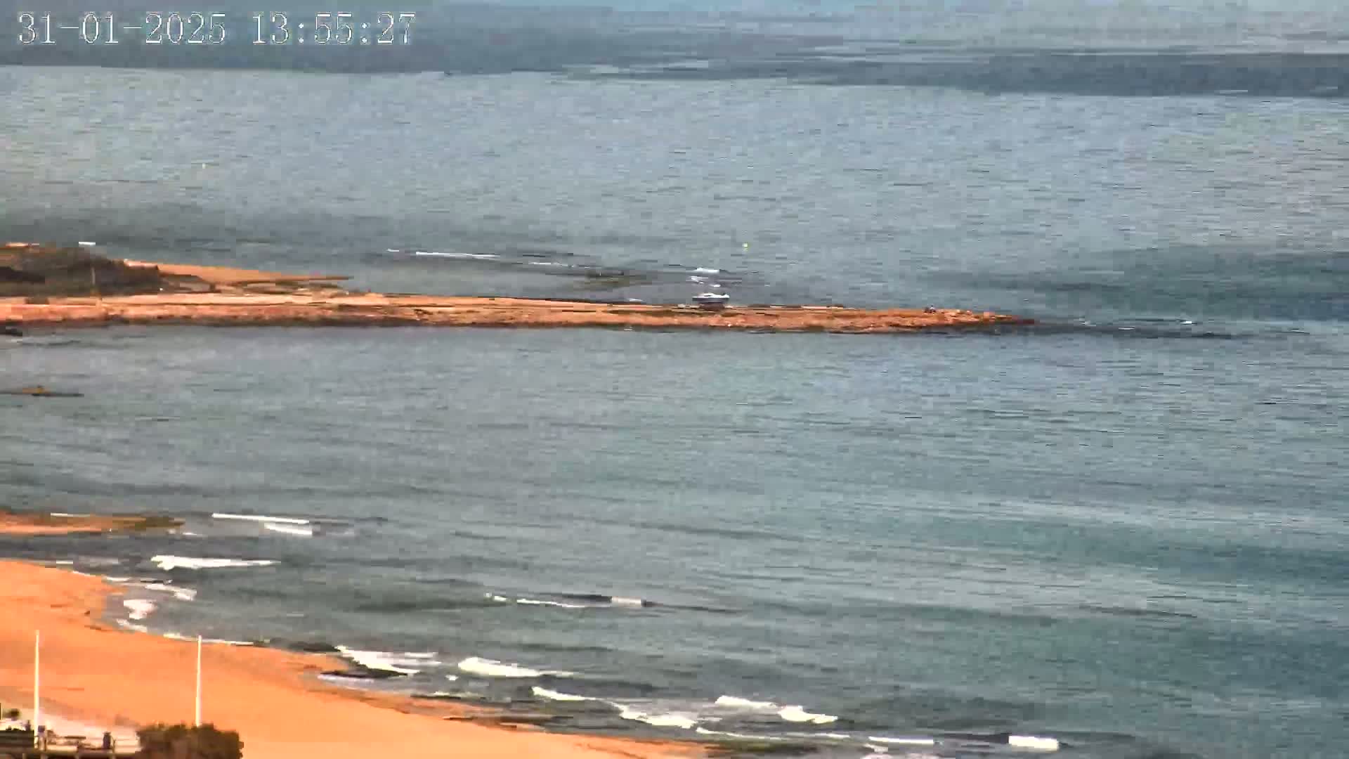 A sandy beach with small waves meets a calm sea where a narrow strip of land extends from the shore into the water.