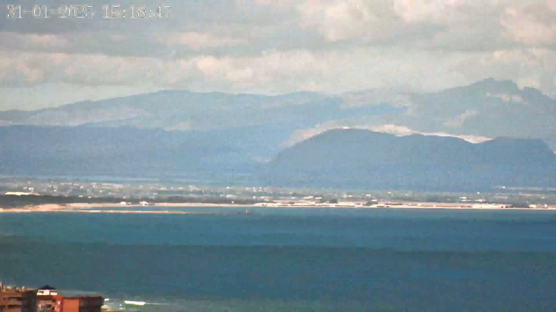 A hazy view shows a body of water stretching to a distant shoreline and mountains under a partly cloudy sky.