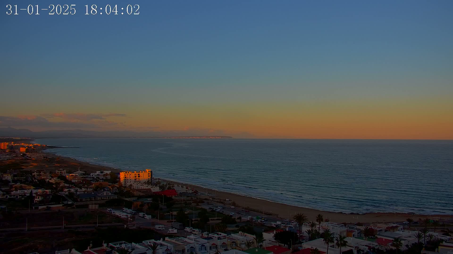 A coastal town at sunset overlooks a calm ocean with a distant landmass under a clear, orange-hued sky.