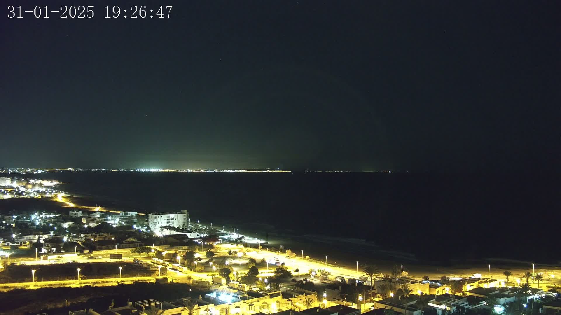 A night-time coastal scene shows a dark ocean under a clear, starry sky, with a brightly lit town stretching along the beach.
