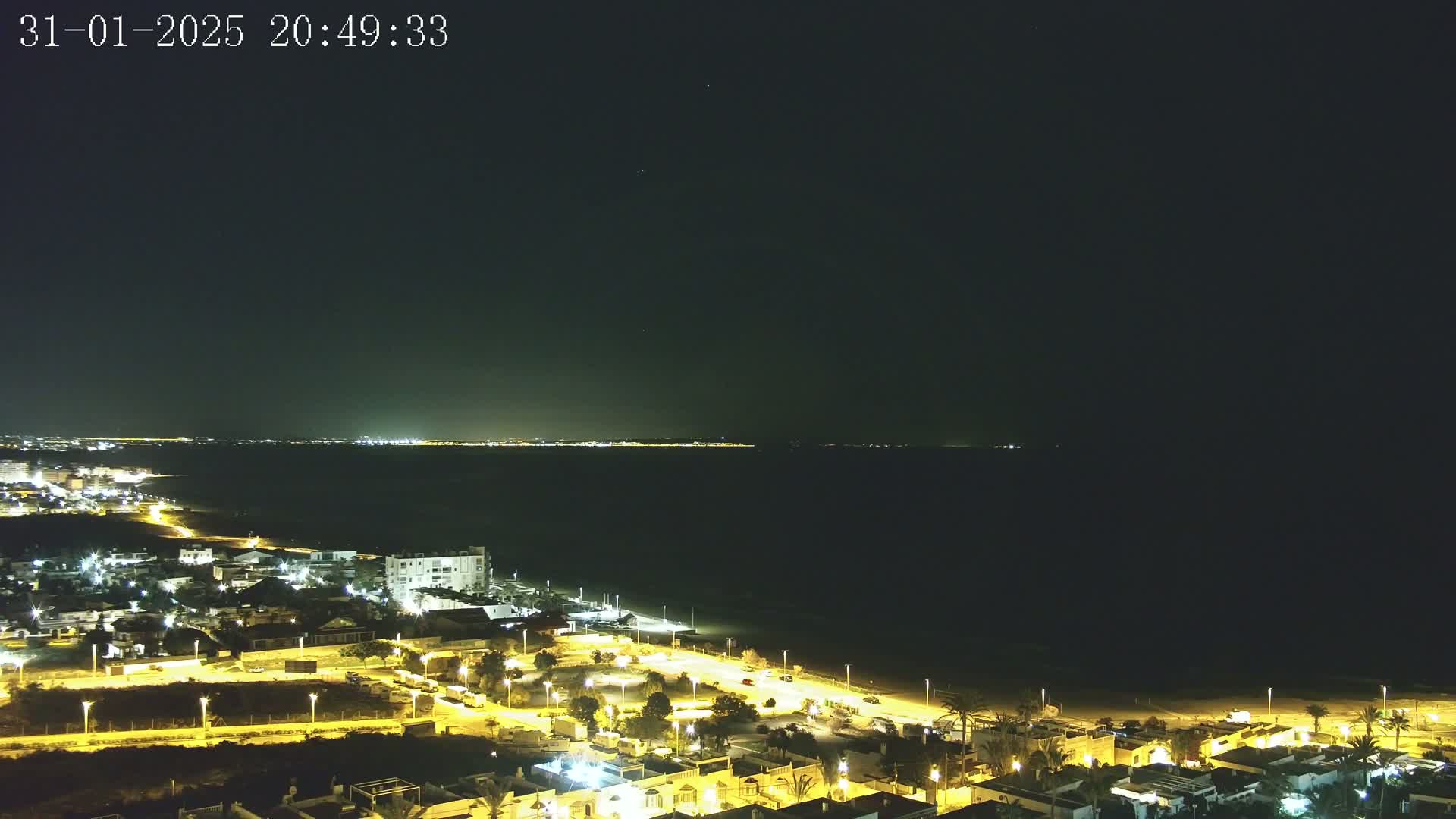 A nighttime aerial view shows a coastal town illuminated by streetlights, bordering a dark sea under a clear night sky.