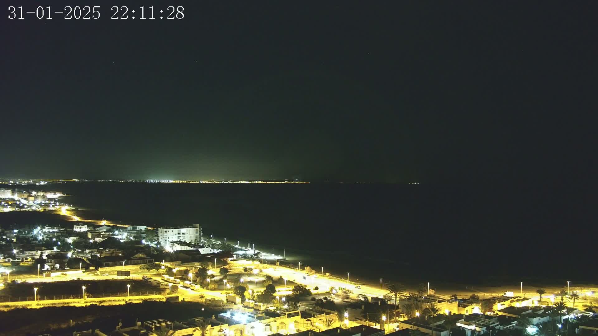 A nighttime, aerial view shows a dark ocean meeting a coastline of illuminated buildings and streets, under a clear, dark sky.