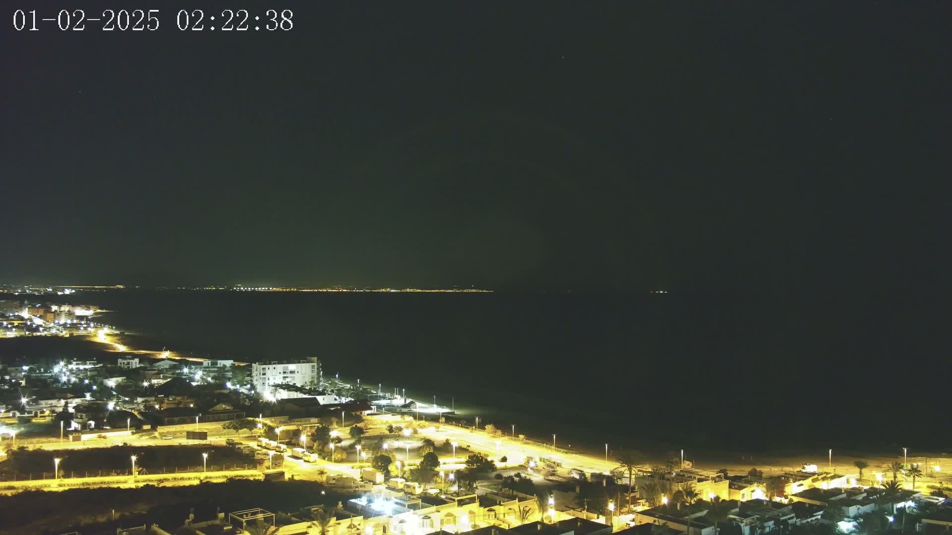 A nighttime aerial view shows a coastal town illuminated by streetlights, with a dark sea extending to a distant, faintly lit shoreline under a clear, star-studded sky.