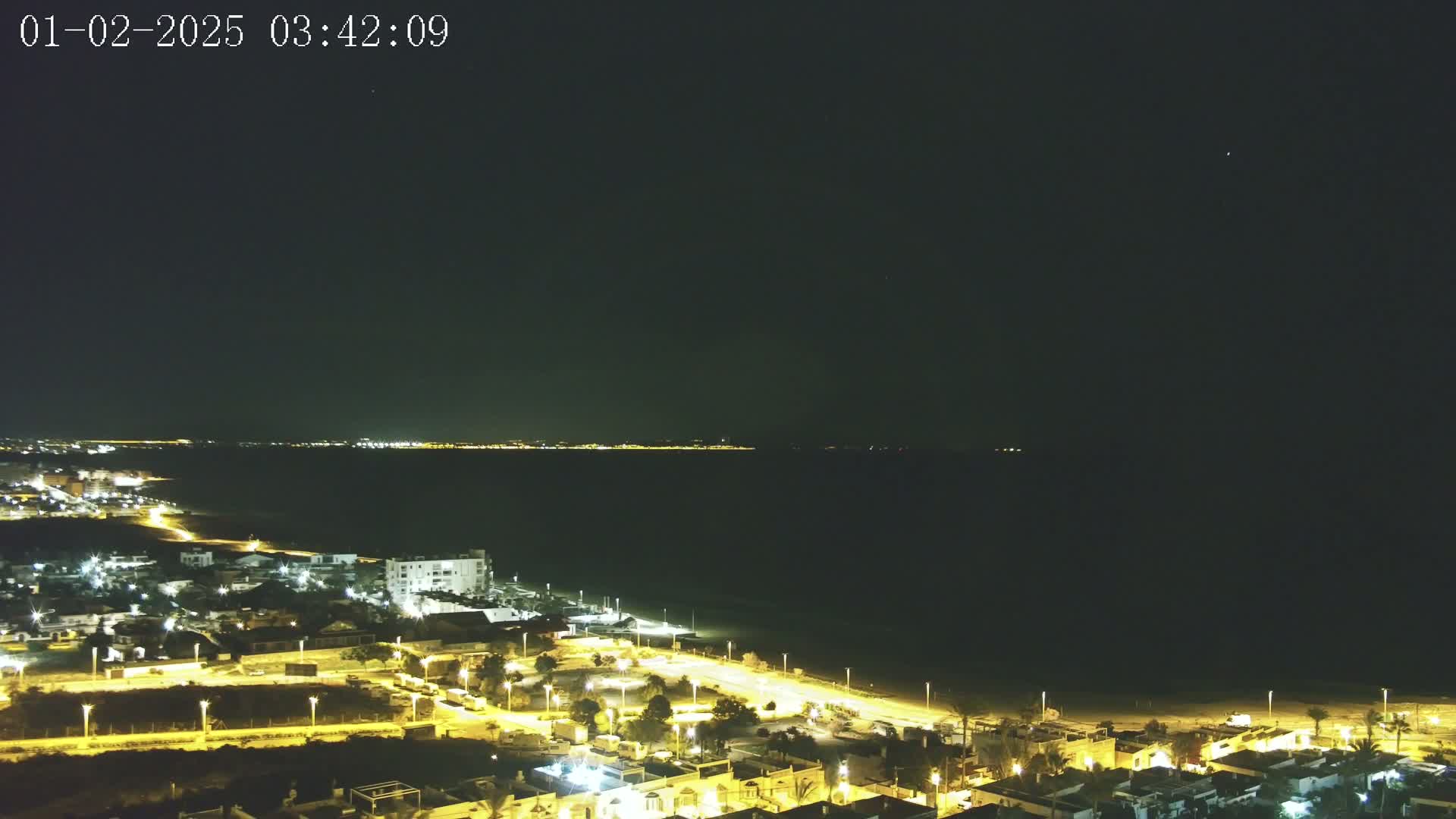 A nighttime aerial view shows a coastal town illuminated by streetlights, overlooking a dark ocean with a distant, faintly lit shoreline under a clear, dark sky.