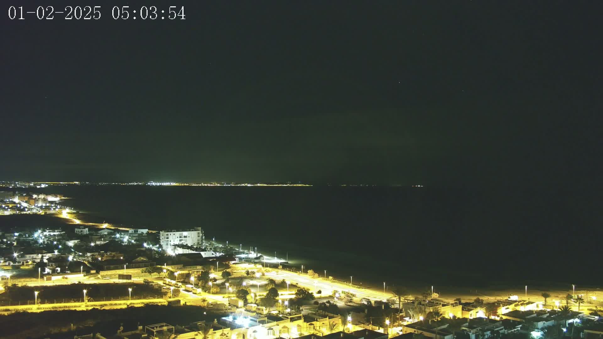 A nighttime coastal scene shows a dark sea under a clear sky, with a town's illuminated streets and buildings stretching along the shoreline and across the land.