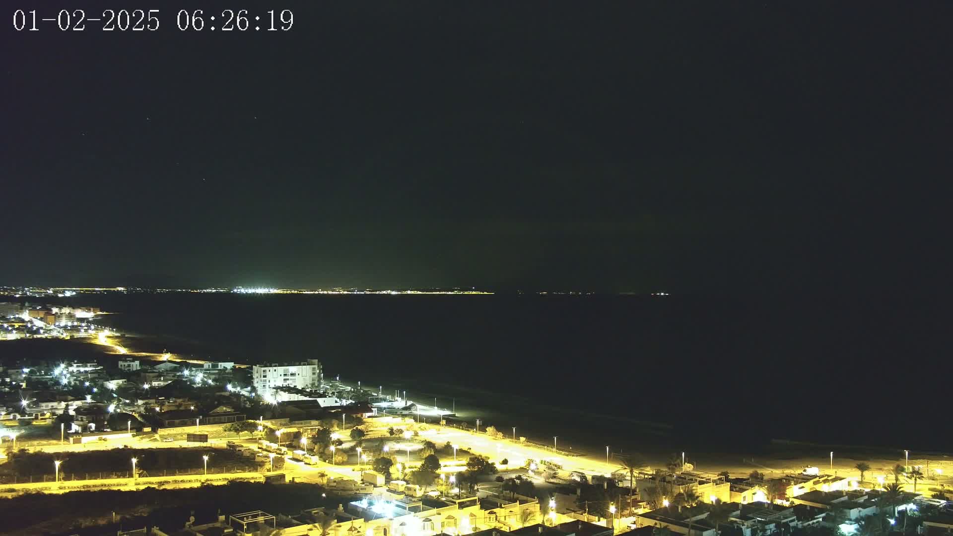 A night-time, high-angle view shows a coastal town illuminated with streetlights, extending to a dark beach and calm sea with a distant, faintly lit shoreline under a clear, dark sky.