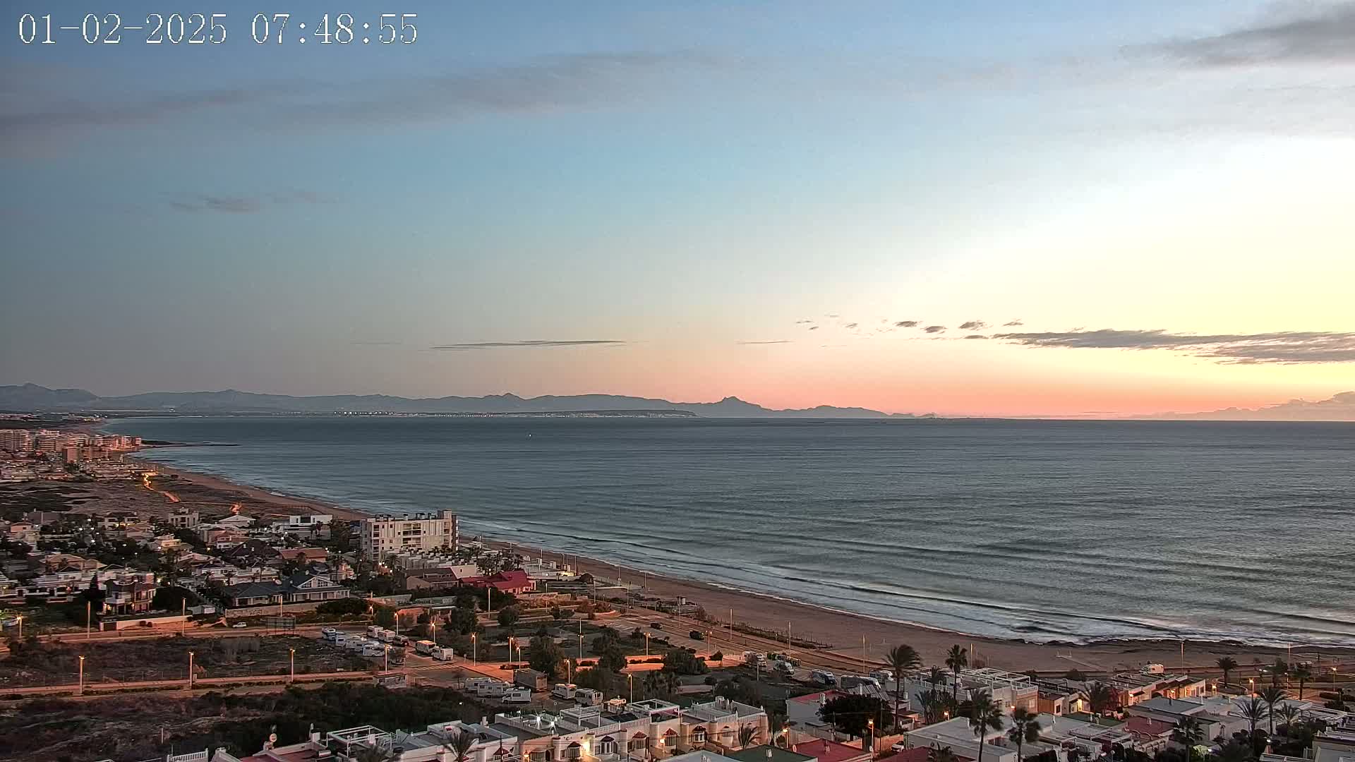 A coastal town at sunset overlooks a calm ocean with gentle waves, a sandy beach, and distant mountains under a pale pink and blue sky.