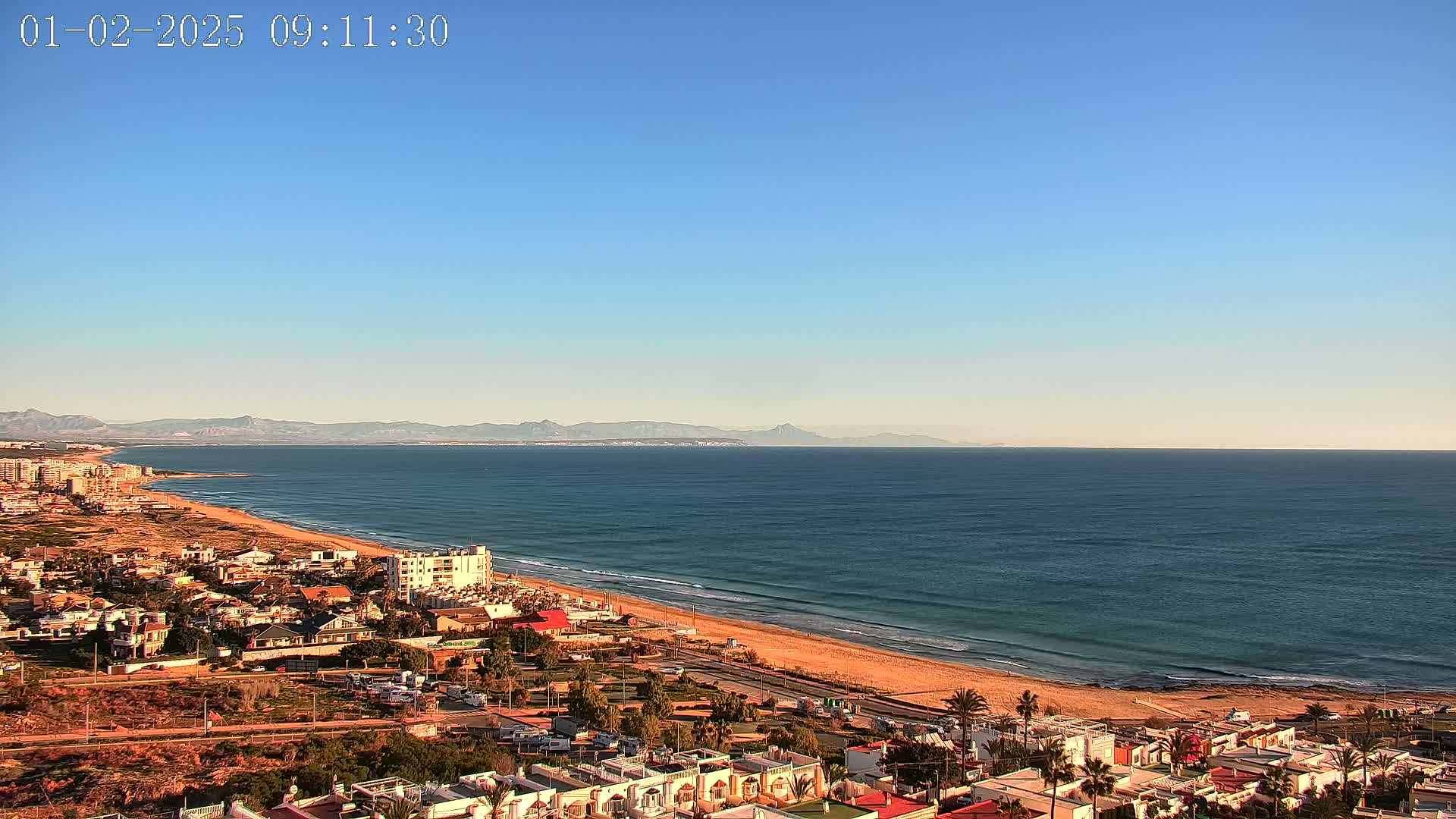 A coastal town overlooks a wide, sandy beach and calm ocean, with mountains visible in the distance under a clear blue sky.