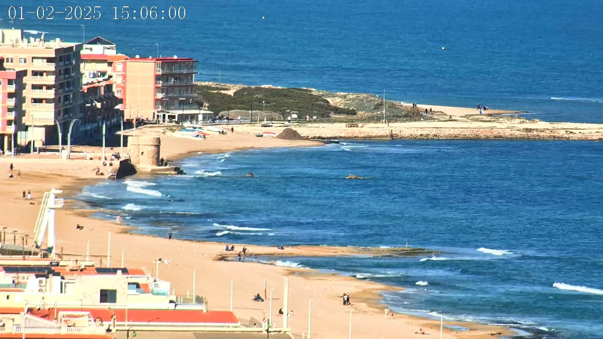 A sandy beach with a few people on it curves around a small, rocky point, with buildings and a stone tower visible in the background under a clear, sunny sky.