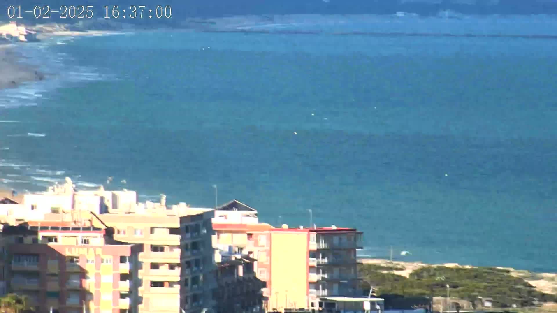 A mostly sunny day shows a teal ocean with small waves lapping a sandy shore where multi-story buildings are visible, with some vegetation on the land.