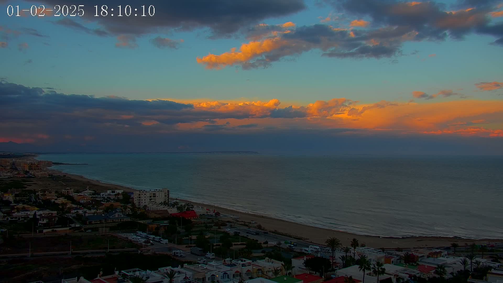 A coastal town at sunset, with partly cloudy skies showing shades of orange and purple, overlooks a calm sea with a sandy beach.
