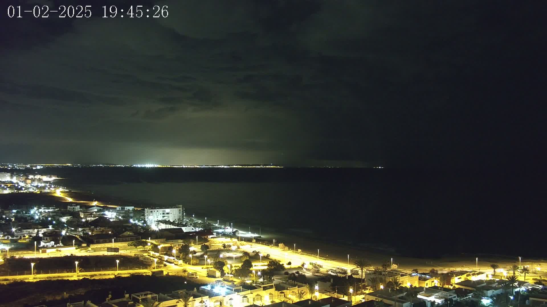 A nighttime coastal town with illuminated streets and buildings overlooks a dark ocean under a cloudy sky.
