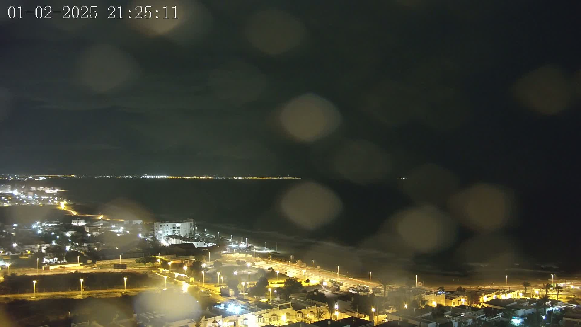 A rain-streaked nighttime view shows a coastal town with illuminated streets and buildings, stretching toward a dark ocean with a faint, distant shoreline of lights.